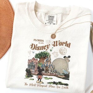 Puede incluir: Camiseta de color crema con un dise&ntilde;o gr&aacute;fico de estilo vintage de Disney World en Florida. El dise&ntilde;o incluye el texto "Florida Disney World" y "The Most Magical Place On Earth", junto con ilustraciones de lugares emblem&aacute;ticos del parque y Mickey Mouse.