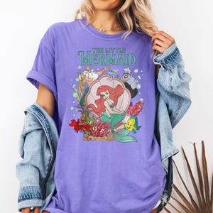 Op de afbeelding: Een lila T-shirt met een afbeelding van "De Kleine Zeemeermin". Het ontwerp bevat Ariel, Botje, Sebastiaan, Ursula en andere personages. De woorden "THE LITTLE MERMAID" staan in groen boven de personages.