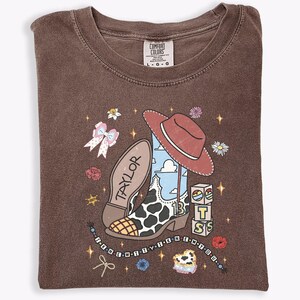 Może przedstawiać: Brązowy t-shirt z okrągłym dekoltem z nadrukiem kowbojskiego buta w łaty, czerwonego kapelusza oraz klock&oacute;w z literami T, S i 13. Napis "TAYLOR" znajduje się na podeszwie buta, a "I KNEW IT I KNEW IT YOU" na sznurku z koralikami.