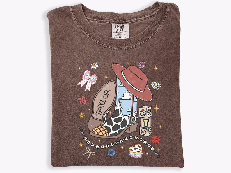 Może przedstawiać: Brązowy t-shirt z okrągłym dekoltem z nadrukiem kowbojskiego buta w łaty, czerwonego kapelusza oraz klock&oacute;w z literami T, S i 13. Napis "TAYLOR" znajduje się na podeszwie buta, a "I KNEW IT I KNEW IT YOU" na sznurku z koralikami.
