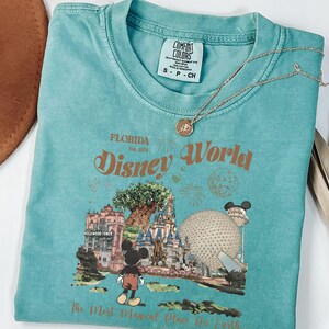 Puede incluir: Una camiseta turquesa Comfort Colors con un gr&aacute;fico de Disney World, que incluye a Mickey Mouse y un castillo, y el texto "The Most Magical Place On Earth". Un collar dorado descansa sobre la camisa. Un sombrero marr&oacute;n es visible en la esquina superior izquierda.