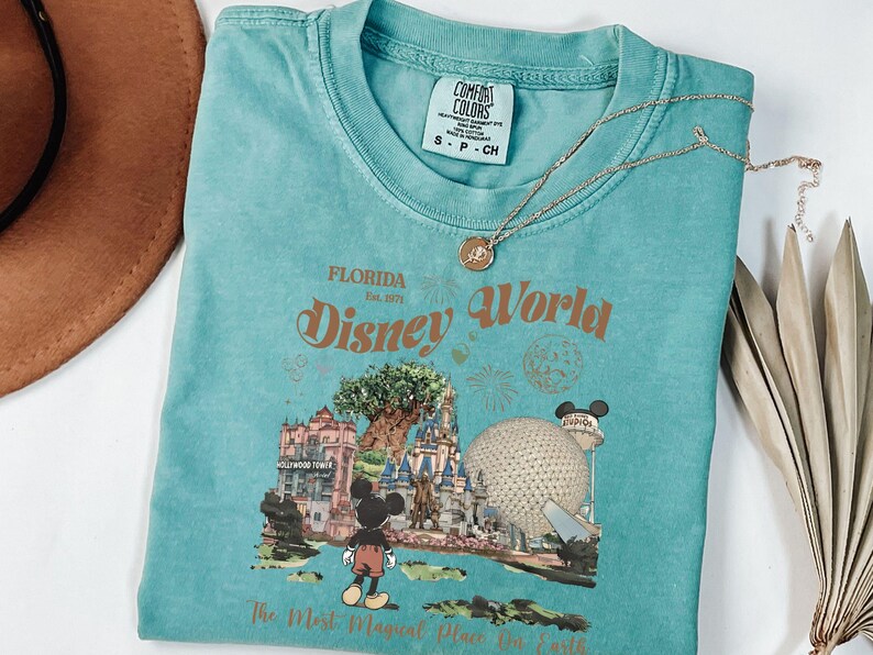 Puede incluir: Una camiseta turquesa Comfort Colors con un gr&aacute;fico de Disney World, que incluye a Mickey Mouse y un castillo, y el texto "The Most Magical Place On Earth". Un collar dorado descansa sobre la camisa. Un sombrero marr&oacute;n es visible en la esquina superior izquierda.