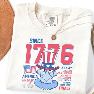 May include: Cream-colored t-shirt featuring a patriotic design with the text "SINCE 1776" in red and blue. The design includes an illustration of Sam Eagle in a Uncle Sam hat, an American flag, and the text "JULY 4TH A SALUTE TO ALL NATIONS BUT MOSTLY AMERICA WITH A GLORIOUS THREE HOUR FINALE."
