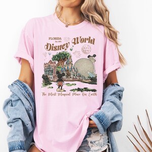 Puede incluir: Camiseta rosa claro con un gr&aacute;fico de Disney World. El dise&ntilde;o incluye el texto "Florida Est. 1971 Disney World" con ilustraciones del &Aacute;rbol de la Vida, Spaceship Earth y Mickey Mouse. Tambi&eacute;n se incluye el texto "The Most Magical Place On Earth".