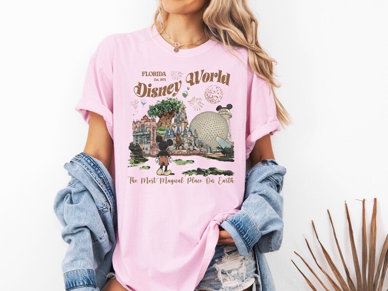 Puede incluir: Camiseta rosa claro con un gr&aacute;fico de Disney World. El dise&ntilde;o incluye el texto "Florida Est. 1971 Disney World" con ilustraciones del &Aacute;rbol de la Vida, Spaceship Earth y Mickey Mouse. Tambi&eacute;n se incluye el texto "The Most Magical Place On Earth".