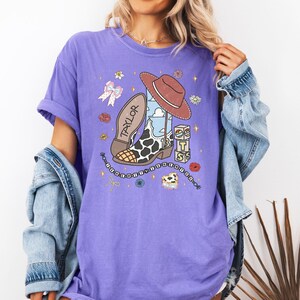 Może przedstawiać: Fioletowy t-shirt z kr&oacute;tkim rękawem z nadrukiem graficznym przedstawiającym brązowy kowbojski kapelusz, but w krowie łaty z imieniem Taylor i numerem 13, łańcuch, kokardę oraz klocki z literami T, S i S, otoczone małymi ikonami kwiat&oacute;w i gwiazd.