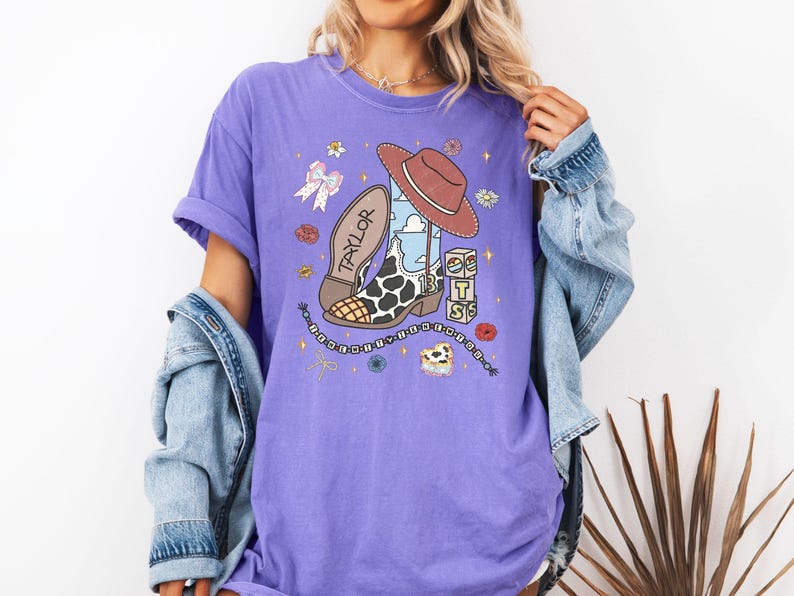 Może przedstawiać: Fioletowy t-shirt z kr&oacute;tkim rękawem z nadrukiem graficznym przedstawiającym brązowy kowbojski kapelusz, but w krowie łaty z imieniem Taylor i numerem 13, łańcuch, kokardę oraz klocki z literami T, S i S, otoczone małymi ikonami kwiat&oacute;w i gwiazd.