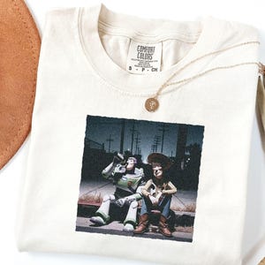 May include: A cream-colored t-shirt with a Comfort Colors label at the neck, featuring a graphic print of Buzz Lightyear and Woody sitting on a curb at night. The shirt is made of 100% cotton and is a size small, as indicated on the interior tag.