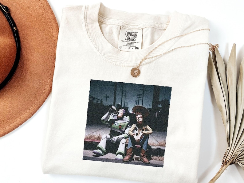 May include: A cream-colored t-shirt with a Comfort Colors label at the neck, featuring a graphic print of Buzz Lightyear and Woody sitting on a curb at night. The shirt is made of 100% cotton and is a size small, as indicated on the interior tag.