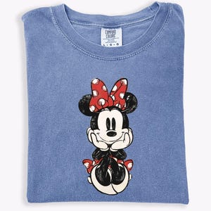 May include: A blue Comfort Colors t-shirt featuring a graphic of Minnie Mouse. Minnie is depicted with a red bow with white polka dots, black ears, and a black outfit. The shirt is folded neatly.