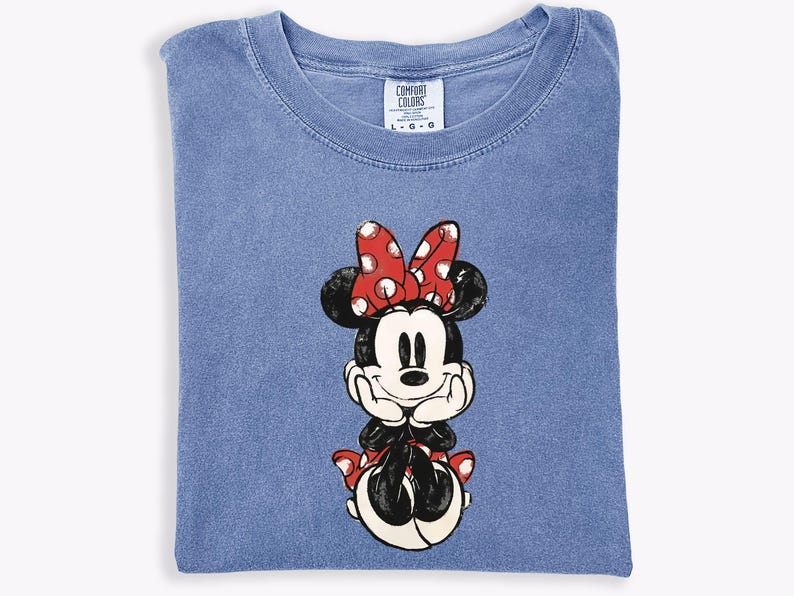 May include: A blue Comfort Colors t-shirt featuring a graphic of Minnie Mouse. Minnie is depicted with a red bow with white polka dots, black ears, and a black outfit. The shirt is folded neatly.