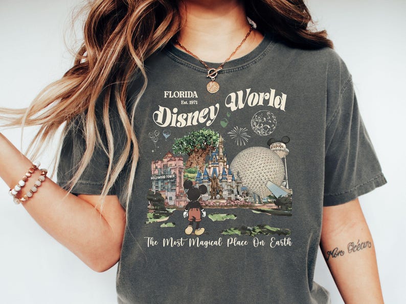 Puede incluir: Camiseta gris oscuro con un dise&ntilde;o vintage de Disney World. El dise&ntilde;o incluye el nombre del parque, la fecha de fundaci&oacute;n y lugares emblem&aacute;ticos como el castillo de Cenicienta y Spaceship Earth, con Mickey Mouse. El texto dice "The Most Magical Place On Earth."