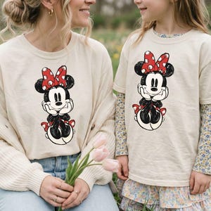 May include: Two beige t-shirts featuring a Minnie Mouse graphic. The graphic shows Minnie Mouse in a seated pose, with a red bow with white polka dots. One shirt is long-sleeved, and the other is short-sleeved.