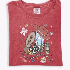 Może przedstawiać: Czerwony t-shirt z grafiką kowbojskiego buta w łaty, brązowym kapeluszem i klockami z literami T, S oraz S. Napis TAYLOR znajduje się na podeszwie buta, a tekst I KNEW YOU WERE TROUBLE jest umieszczony na sznurku z koralikami.