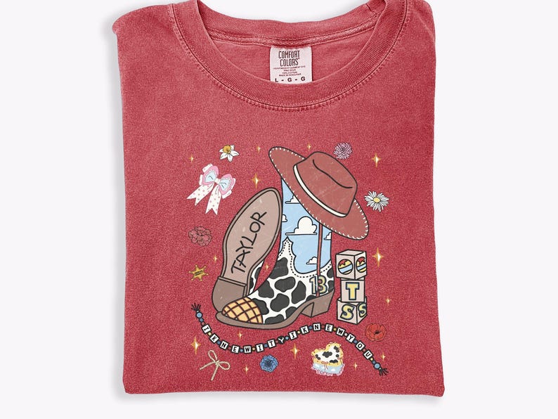 Może przedstawiać: Czerwony t-shirt z grafiką kowbojskiego buta w łaty, brązowym kapeluszem i klockami z literami T, S oraz S. Napis TAYLOR znajduje się na podeszwie buta, a tekst I KNEW YOU WERE TROUBLE jest umieszczony na sznurku z koralikami.