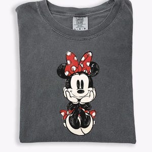 May include: Dark gray Comfort Colors t-shirt featuring a distressed Minnie Mouse graphic. Minnie is depicted in black, white, and red, with a polka-dot bow. The shirt is folded and has a tag in the upper center.