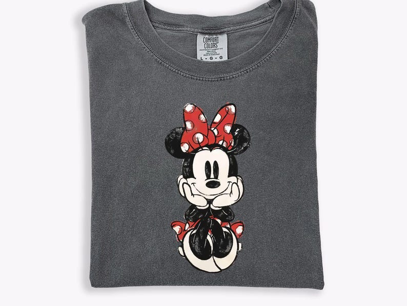 May include: Dark gray Comfort Colors t-shirt featuring a distressed Minnie Mouse graphic. Minnie is depicted in black, white, and red, with a polka-dot bow. The shirt is folded and has a tag in the upper center.