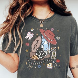 Może przedstawiać: Ciemnoszary t-shirt z nadrukiem graficznym przedstawiającym kowbojski but w łaty, brązowy kapelusz, imię TAYLOR, liczbę 13, klocki z literami T S, kokardę, kwiaty, gwiazdki oraz sznurek z koralikami i napisem.