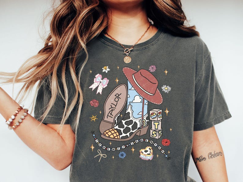 Może przedstawiać: Ciemnoszary t-shirt z nadrukiem graficznym przedstawiającym kowbojski but w łaty, brązowy kapelusz, imię TAYLOR, liczbę 13, klocki z literami T S, kokardę, kwiaty, gwiazdki oraz sznurek z koralikami i napisem.