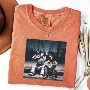 May include: A terracotta-colored short-sleeve t-shirt featuring a graphic print of two animated characters sitting on a curb at night. The shirt has a crew neckline and a Comfort Colors brand label on the inside collar. The fabric has a washed texture.
