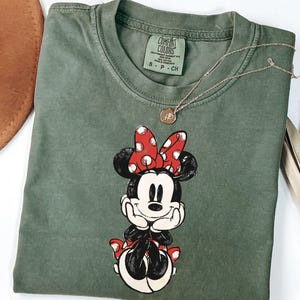 May include: An olive green t-shirt featuring a graphic of Minnie Mouse with a red polka dot bow. The shirt has a crew neck and short sleeves. A gold necklace with a pendant is draped across the shirt. The shirt is folded on a white surface.