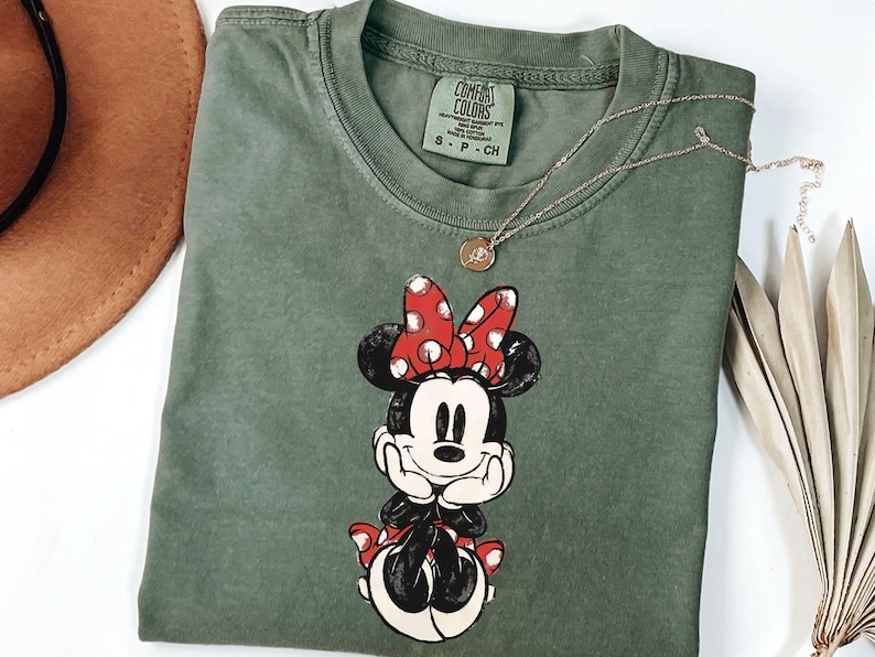 May include: An olive green t-shirt featuring a graphic of Minnie Mouse with a red polka dot bow. The shirt has a crew neck and short sleeves. A gold necklace with a pendant is draped across the shirt. The shirt is folded on a white surface.