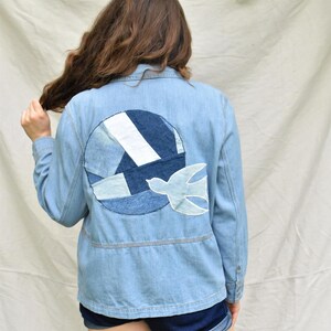 May include: Light blue denim jacket with a circular patch on the back featuring a patchwork design in shades of blue and white, with a light blue bird applique. The jacket has a button-up front and a cinched waist.