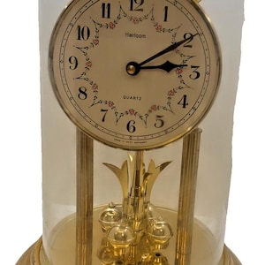 May include: A gold-tone quartz anniversary clock with a glass dome, featuring a white face with black Arabic numerals, floral accents, and the text Heirloom and QUARTZ. The base and internal pendulum structure are made of polished gold-tone metal.