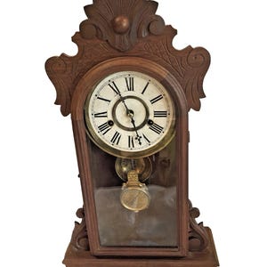 May include: A brown wooden mantel clock featuring an ornate carved top, a circular white face with black Roman numerals, and black hands. A glass panel below the clock face reveals a gold-colored pendulum swinging inside the wooden frame.