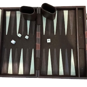 May include: A brown backgammon board game set featuring a dark brown playing surface with alternating light and dark brown triangular points. The set includes white and brown checkers, two dice cups, two standard dice, and one doubling cube with 64.