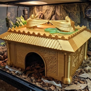 Könnte beinhalten: Ein goldfarbenes, rechteckiges Reptilienversteck in Form eines antiken Tempels mit einem zentralen Bogeneingang. Das Dach zeigt zwei symmetrische, geflügelte Figuren, die sich gegenüberstehen, mit einer grünen Schlange auf der flachen Oberseite.