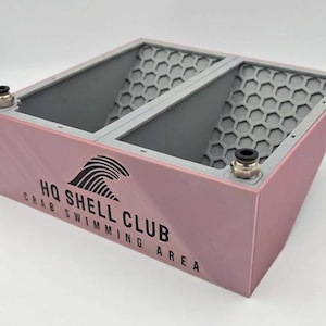 May include: A pink rectangular container with two open compartments featuring grey honeycomb-patterned interior walls. The front panel displays the black text HQ SHELL CLUB CRAB SWIMMING AREA with a wave icon. Two metallic pneumatic fittings are attached.