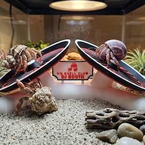 May include: A white plastic DJ booth toy for hermit crabs featuring two black and red circular platforms. The center sign displays the text "HQ SHELL CLUB DJ BOOTH" with a small red crab icon. Three hermit crabs are positioned on and near the structure.