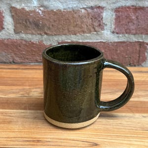 Peut inclure: Une tasse en céramique faite à la main avec un glaçage vert foncé et une base naturelle non émaillée. La tasse a une anse arrondie et une forme cylindrique, idéale pour le café ou le thé. La tasse mesure environ 10 cm de haut.