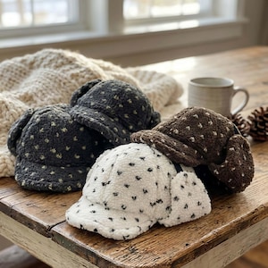 May include: Four fleece hats in shades of gray, brown, and white, each adorned with a repeating pattern of small, dark-colored shapes. The hats have ear flaps and visors, resting on a wooden table. A cozy, cream-colored blanket is in the background.