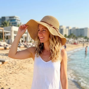 May include: A wide-brimmed straw hat in a natural tan color featuring a woven texture and a thin brown cord tied in a small bow around the base of the crown. The hat has a flexible, downward-sloping brim and a rounded top crown structure.