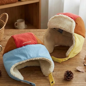 May include: Two colourful trapper hats with ear flaps. One hat is red, brown, and blue, with the text "BOBO POIONE". The other hat is red, brown, and yellow. Both hats have a soft, cream-coloured lining and yellow straps.