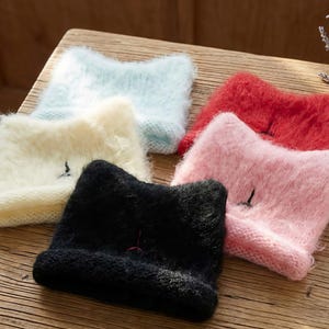 May include: A selection of cat ear beanies in black, pink, red, pale blue, and cream. Each beanie features a small embroidered detail and is made from a soft, fluffy material. The beanies are arranged on a wooden surface.