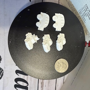 May include: Five small white molded shapes resembling vehicles, including two truck-like forms and three car-like forms, arranged on a black circular surface next to a silver United States quarter coin for scale comparison.