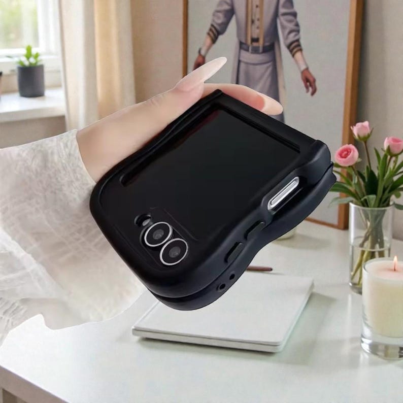 May include: A black phone case with a dual-lens camera cutout. The case has a sleek, curved design and appears to be made of a matte material. The phone case is held in front of a white surface with a painting and flowers in the background.