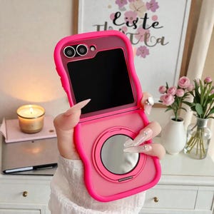 May include: A bright pink phone case with a built-in mirror and camera cutouts. The case is open, revealing the phone's screen. The design includes a ring for a finger grip. The case is held by a person.