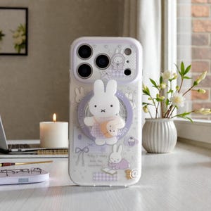 Op de afbeelding: Een doorzichtige telefoonhoes met een wit en paars ontwerp met een cartoon konijn karakter. De hoes heeft een pop-up grip met het konijn en een maan. De hoes heeft ook de tekst "Miffy's story book has everything".