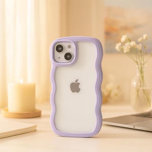 Puede incluir: Funda transparente para iPhone con borde ondulado lila. La funda tiene una parte trasera transparente con el logotipo de Apple. La funda del teléfono está sobre una superficie blanca con una vela, un cuaderno y un portátil en el fondo.
