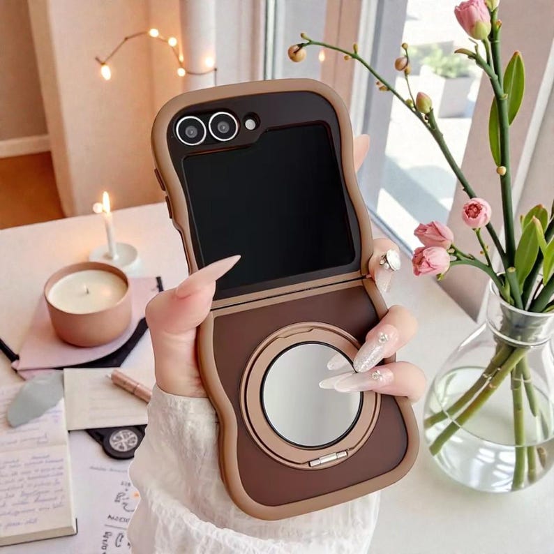 May include: A brown and tan phone case with a built-in mirror. The case has a rectangular shape with rounded edges and a camera cutout. The mirror is round and has a metallic frame. The phone case is designed to protect the phone.