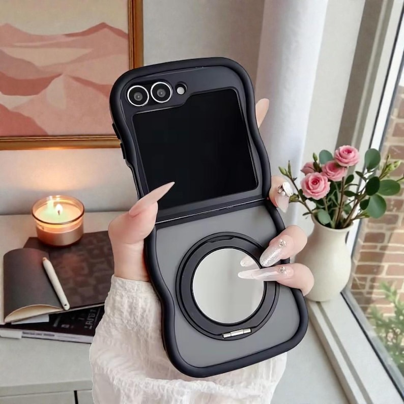 May include: A black and gray phone case with a flip-open design. The case features a circular mirror and a camera cutout. The phone case is held in a person's hand, with a candle and flowers in the background.