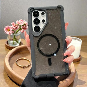 May include: A dark grey phone case with a glittery, textured back and a black circular design. The case has a protective bumper and a camera cutout. The phone case is shown with a gold bracelet and a small vase of pink flowers.