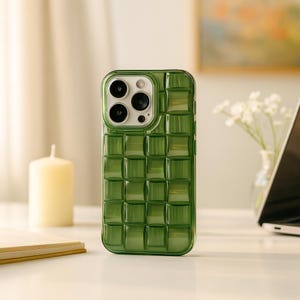 May include: A green phone case with a woven pattern. The case has a square grid design and a clear, glossy finish. The phone case is on a white surface with a candle and a laptop in the background.