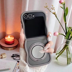 May include: A gray phone case with a circular design on the bottom half, and a camera lens design on the top. The phone case is open, revealing a black screen. The case is held in front of a soft background with a candle and flowers.
