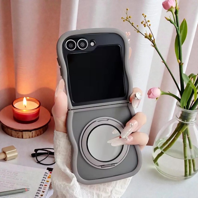 May include: A gray phone case with a circular design on the bottom half, and a camera lens design on the top. The phone case is open, revealing a black screen. The case is held in front of a soft background with a candle and flowers.
