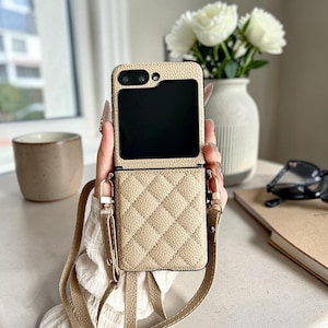 Può includere: Una custodia per smartphone pieghevole, testurizzata e color beige, caratterizzata da un motivo trapuntato sulla metà inferiore e da un ritaglio rettangolare nero liscio per lo schermo. Include una tracolla beige abbinata con finiture dorate.